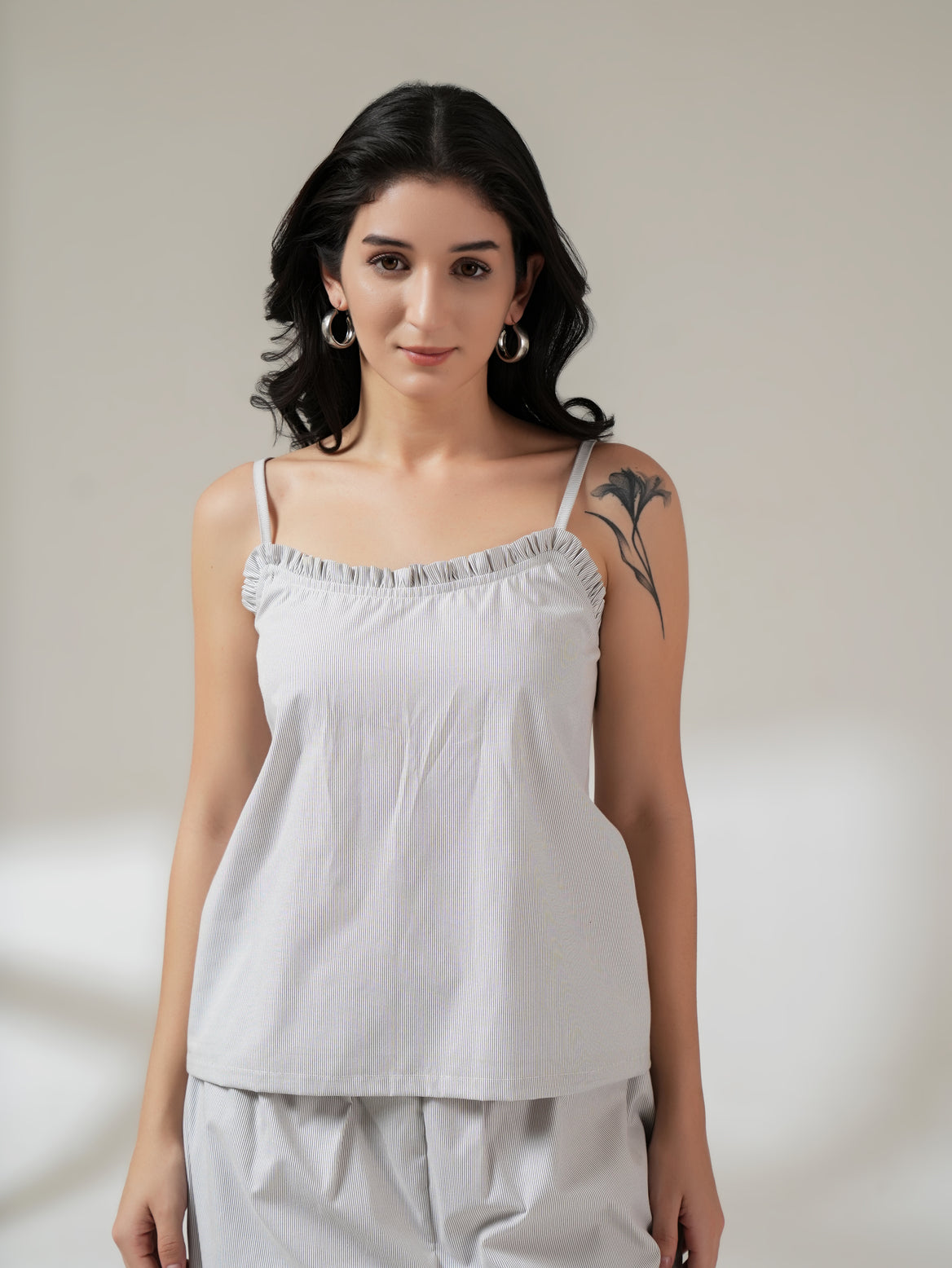 A woman modeling a white and gray vertically striped camisole top with ruffled straps, paired with matching light gray pants, against a neutral beige studio background.