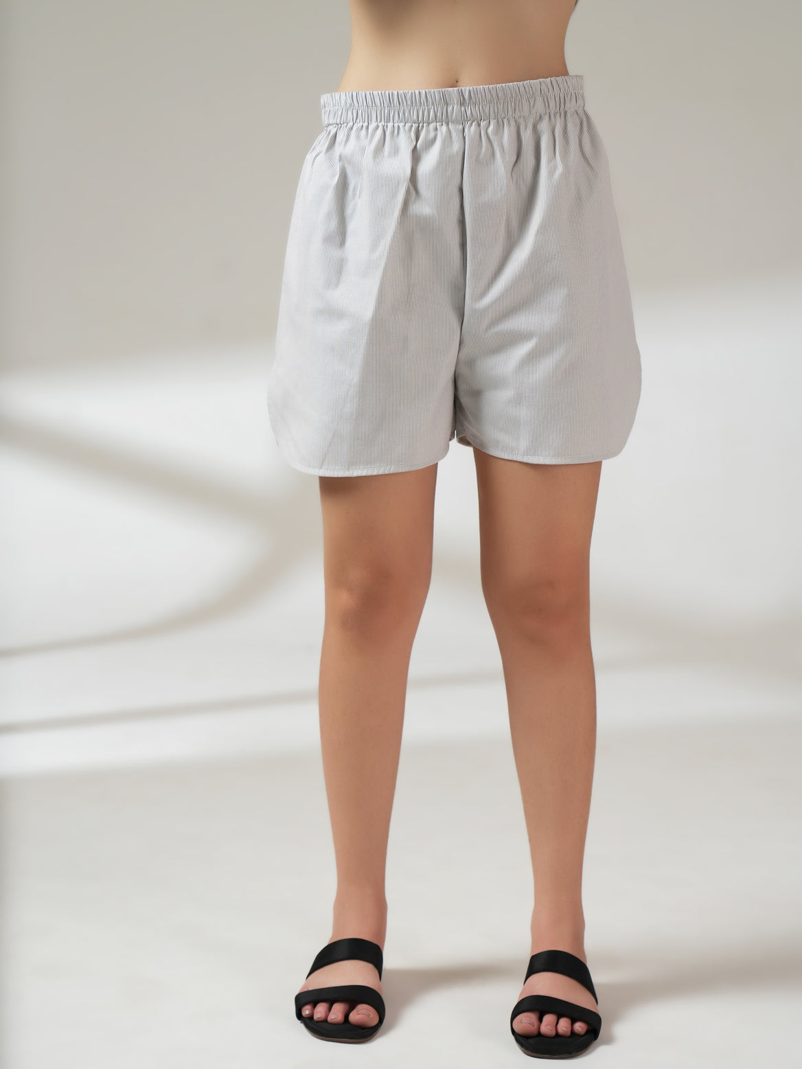 A person wearing light gray and white vertically striped shorts with an elastic waistband, paired with black slide sandals, shown from the waist down against a neutral studio background.