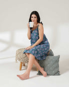 Woman in a blue floral dress holding a mug, sitting on a white surface with a gray pillow.