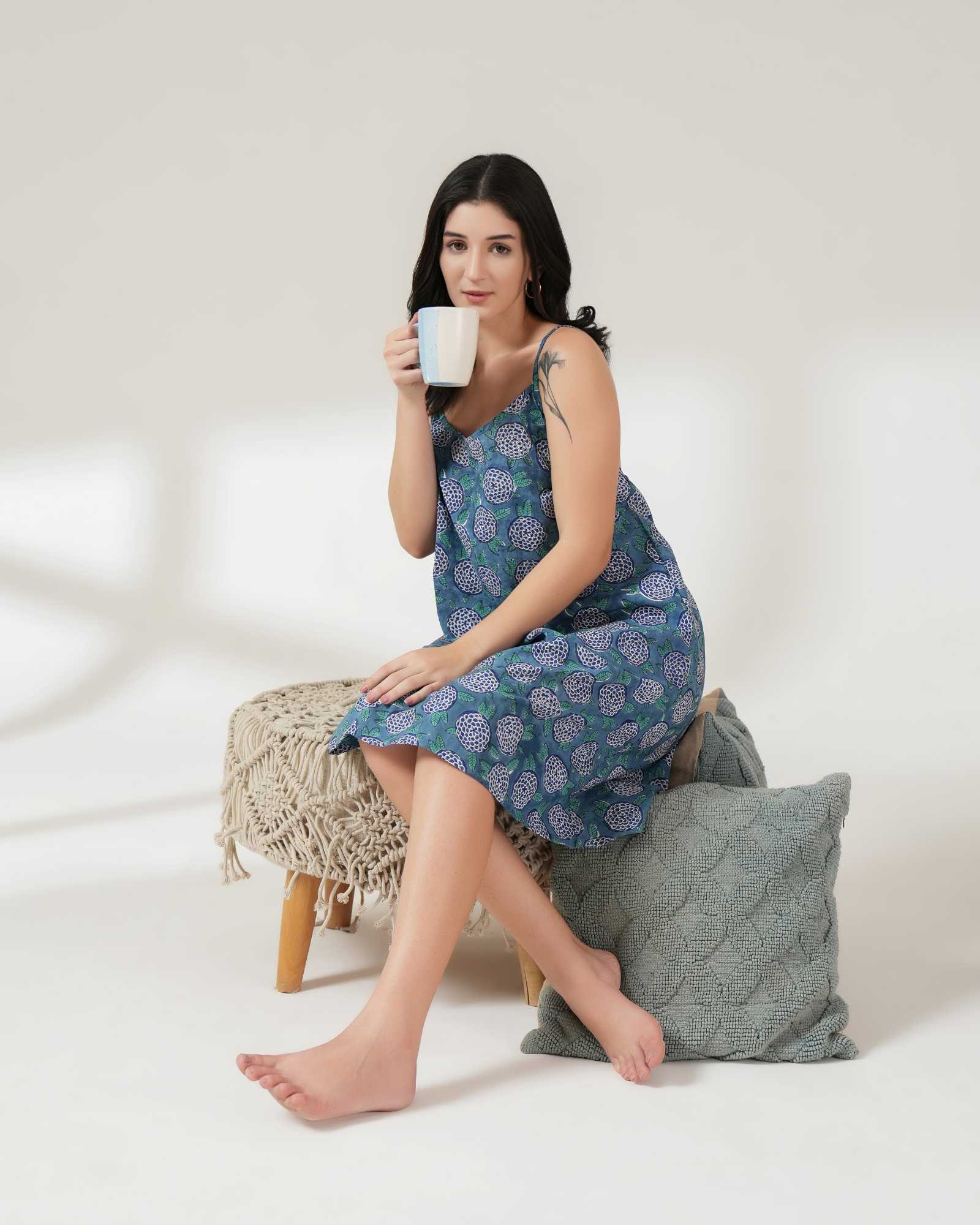 Woman in a blue floral dress holding a mug, sitting on a white surface with a gray pillow.