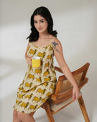 Woman in a yellow and white dress with animal pattern, holding a yellow mug, sitting on a wooden chair.