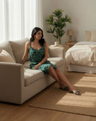 Woman in a green dress sitting on a beige couch in a well-lit room with plants and furniture.