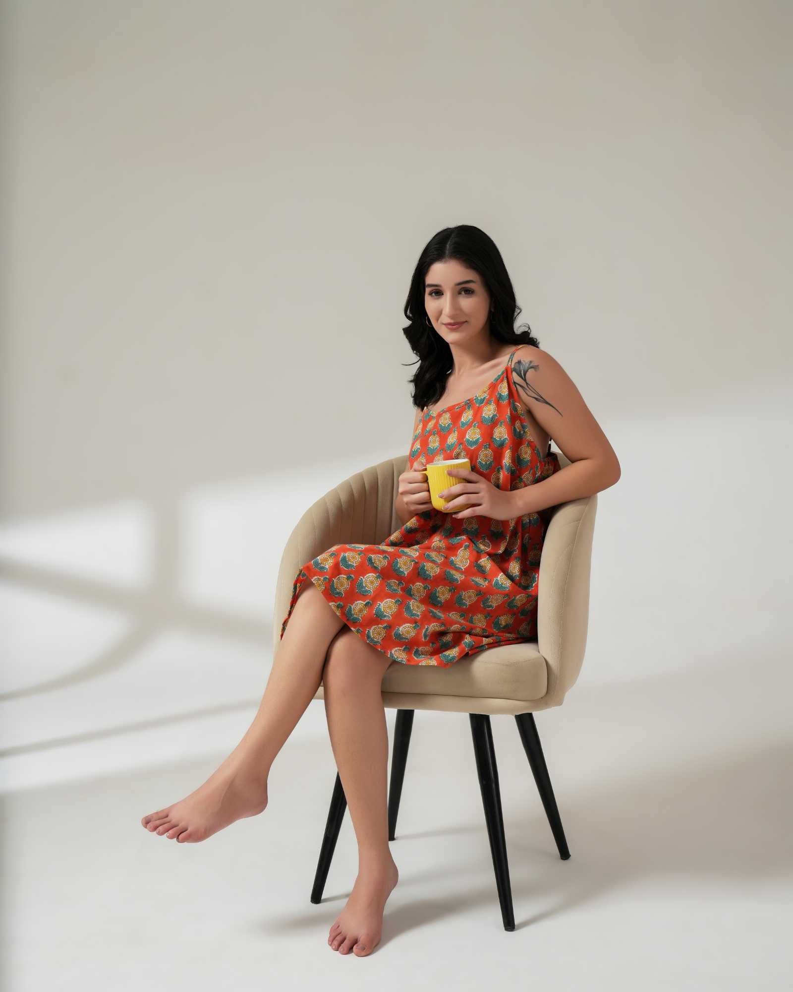 Woman in a patterned dress sitting on a chair holding a yellow cup against a white background