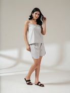 A woman modeling a light gray two-piece lounge set consisting of a sleeveless camisole top and matching shorts, paired with black slide sandals, set against a warm beige studio background with soft window shadow projections.