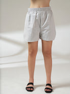 A person wearing light gray and white vertically striped shorts with an elastic waistband, paired with black slide sandals, shown from the waist down against a neutral studio background.